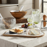 A styled tabletop serving scene with a rectangular wooden board holding crackers and cheese under a clear glass dome, alongside a glass pitcher, glass bowl, wooden bowl, and fruit on a kitchen counter.